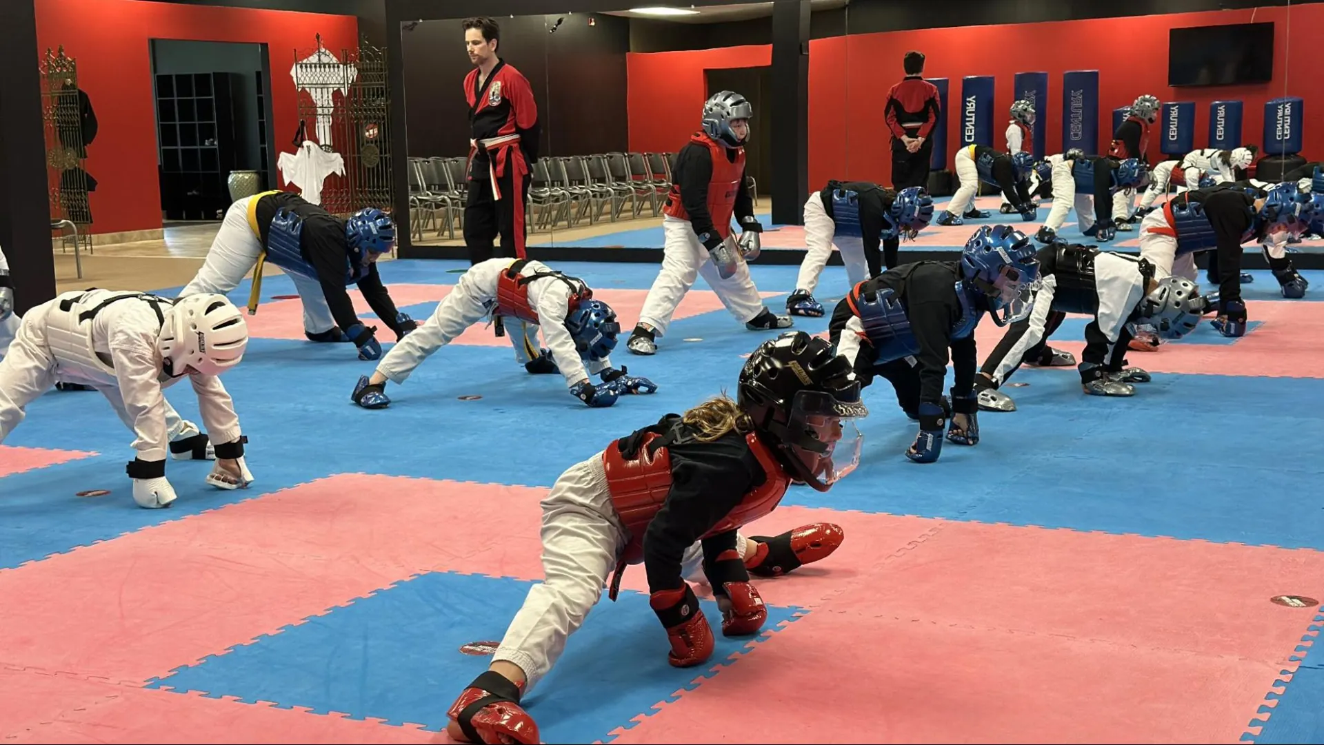 Children in sparring gear stretching on the mat as an instructor supervises at Murray's Martial Arts